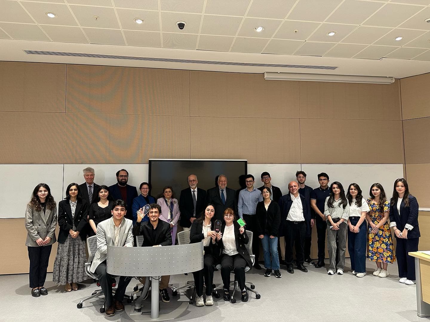 A large group gathered for a photograph at the front of a lecture hall, some seated at the front desk, a dark screen on the wall behind them.