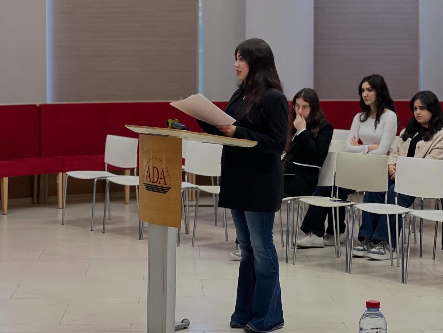 A debater speaking from notes at an ADA lectern, with other participants seated along the red wall behind.