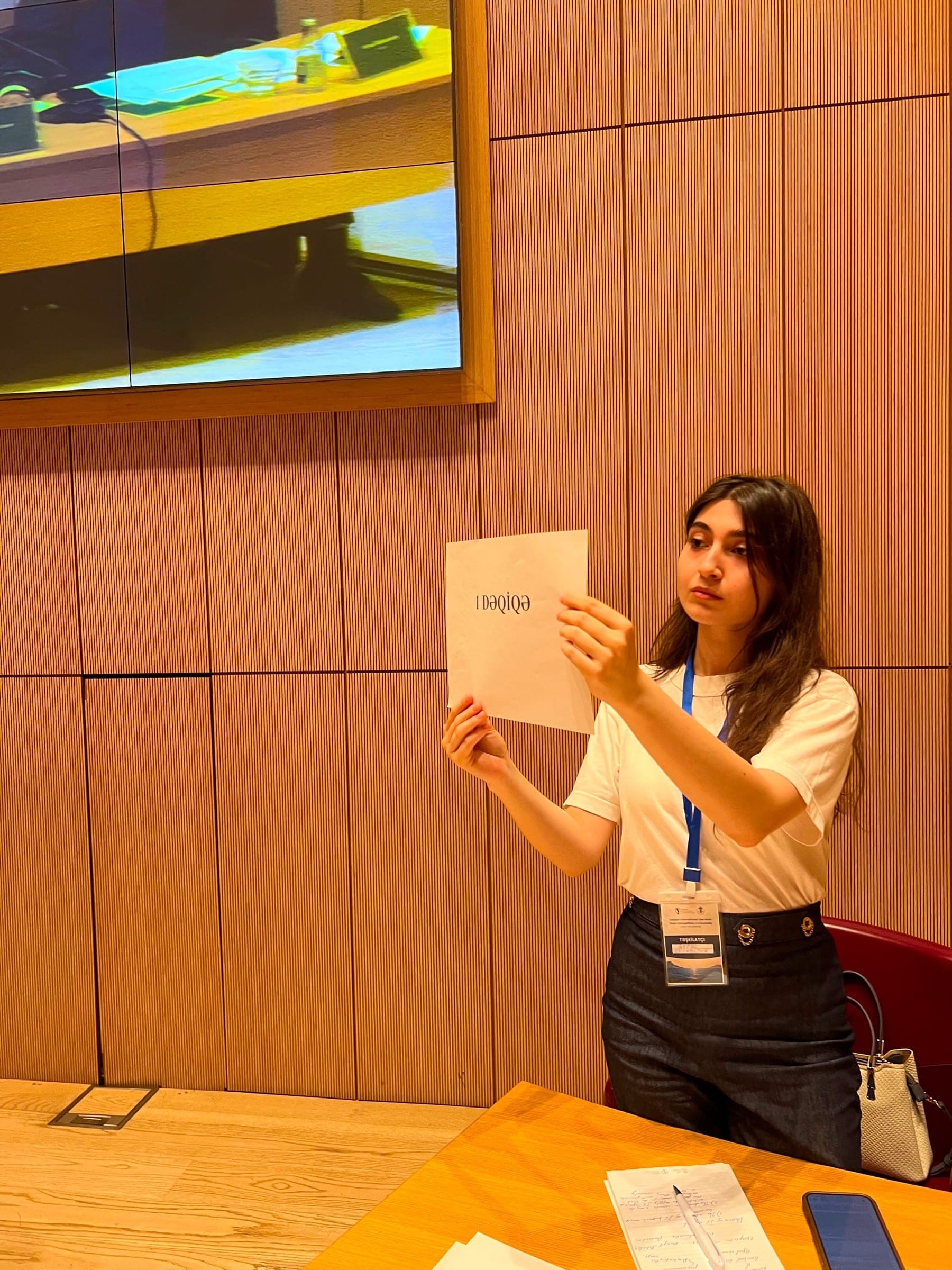 A participant holding up a certificate in the chamber, with a screen showing the bench behind her.
