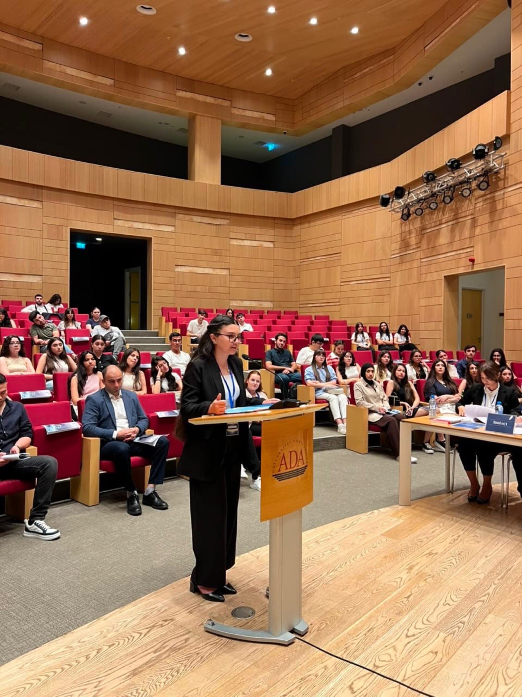 A participant standing at the ADA lectern addressing the court, with the judges seated to one side and the audience behind.