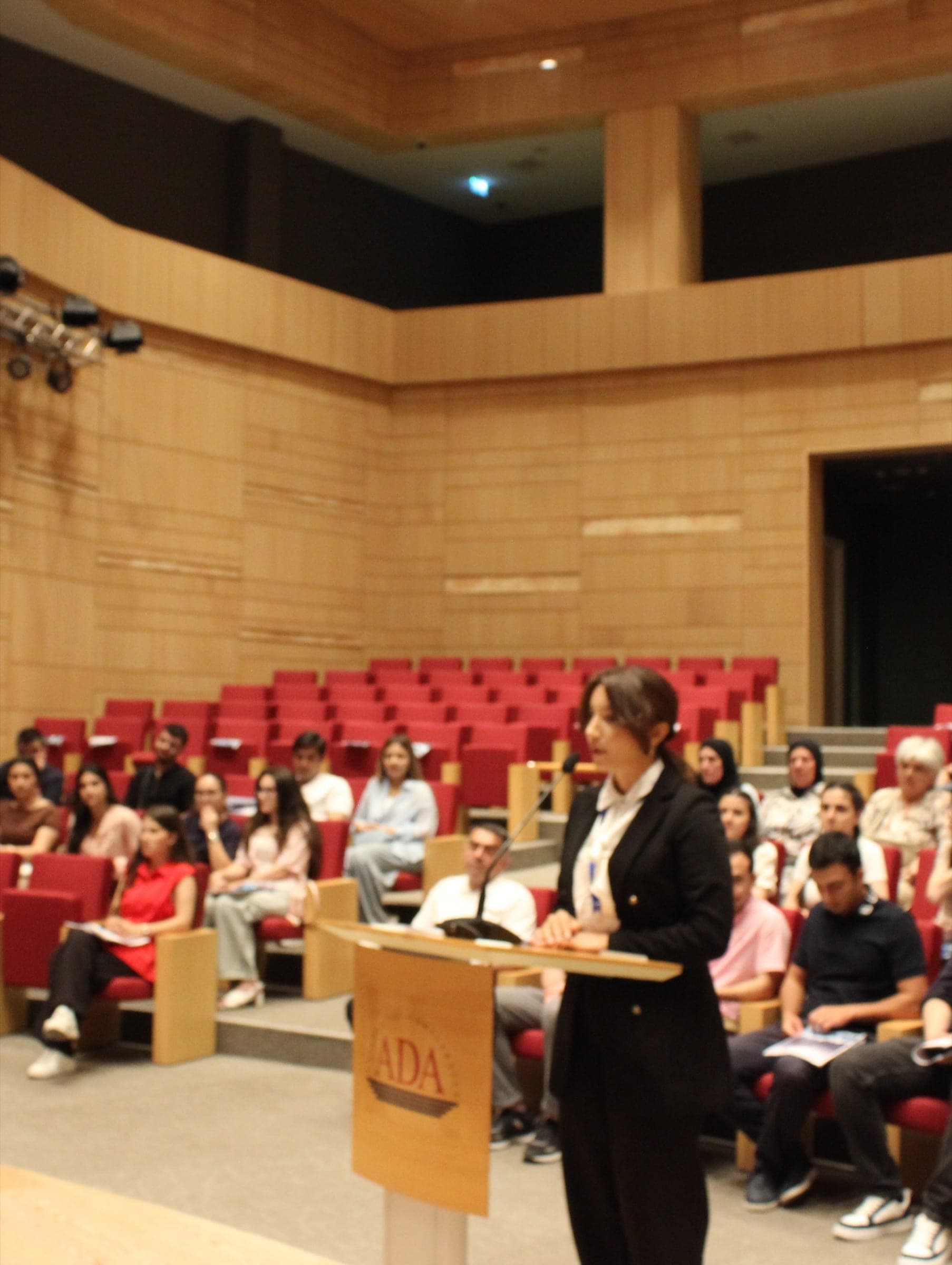 A student speaking from a lectern marked ADA in a wood-panelled auditorium, with rows of red seats filled with listeners behind.