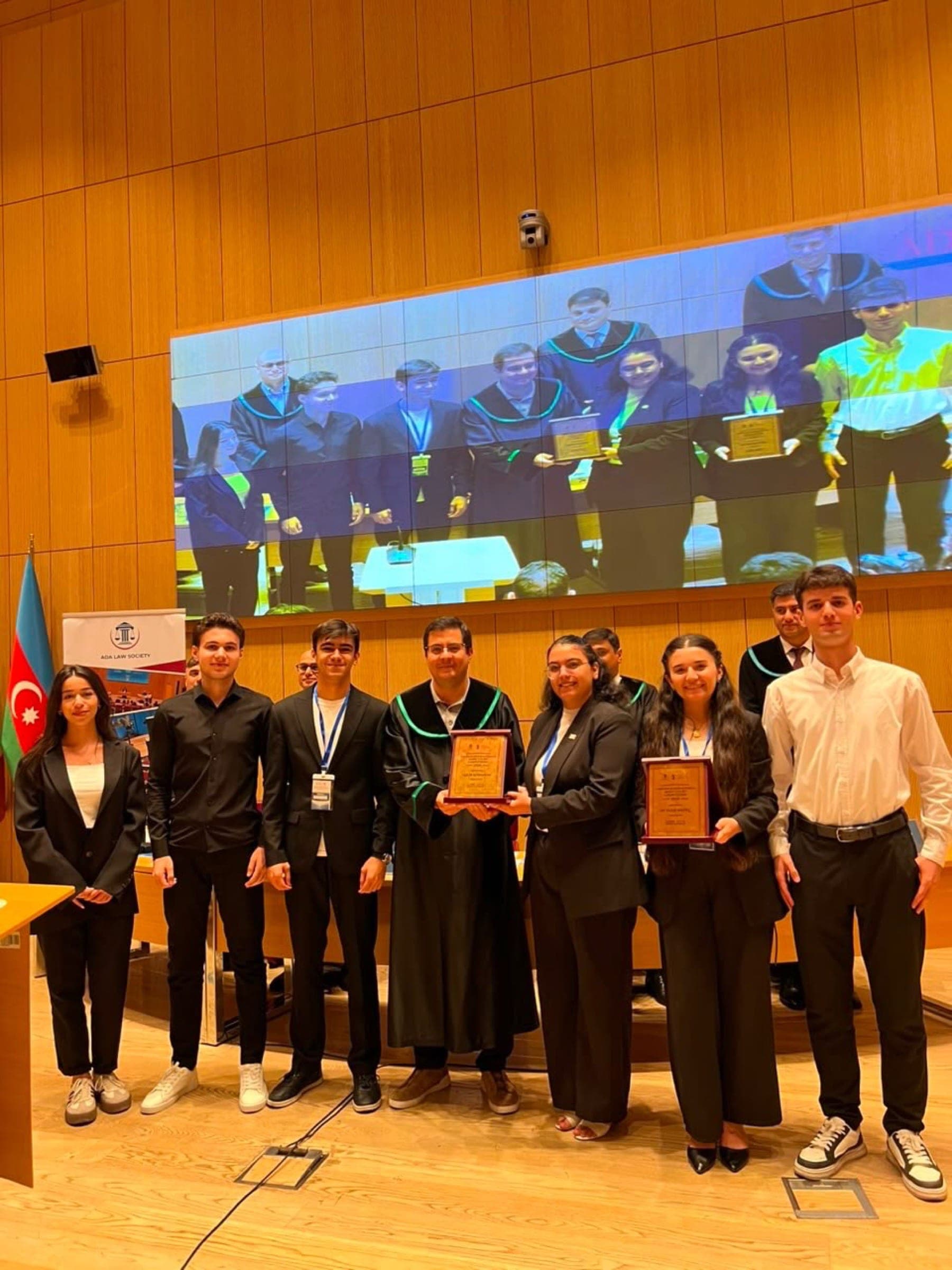 A group of participants holding plaques beside a judge in robes, with the flag of Azerbaijan at the side of the chamber.