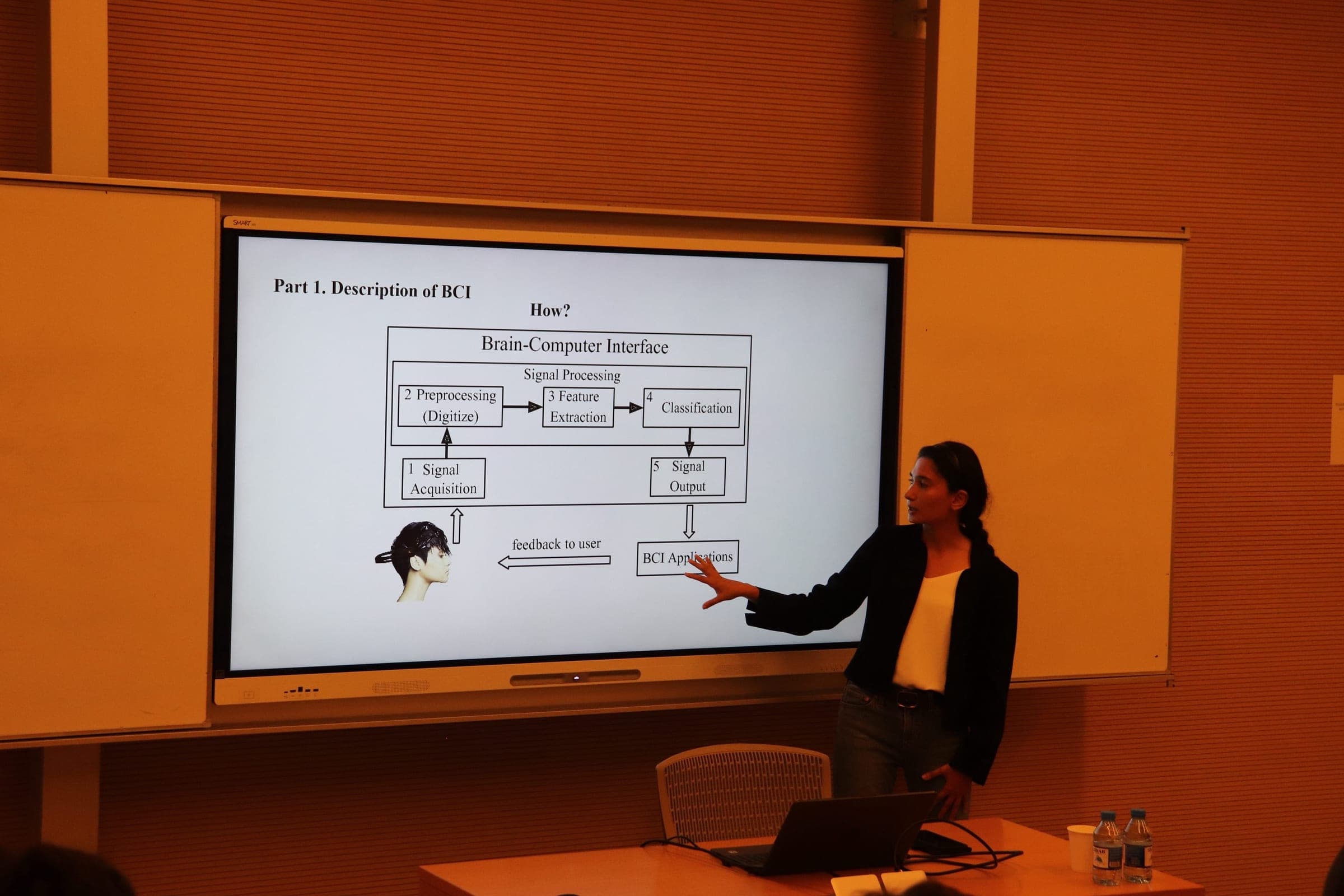The speaker pointing to a diagram of a brain-computer interface on a large screen, under the heading “Part 1. Description of BCI”.