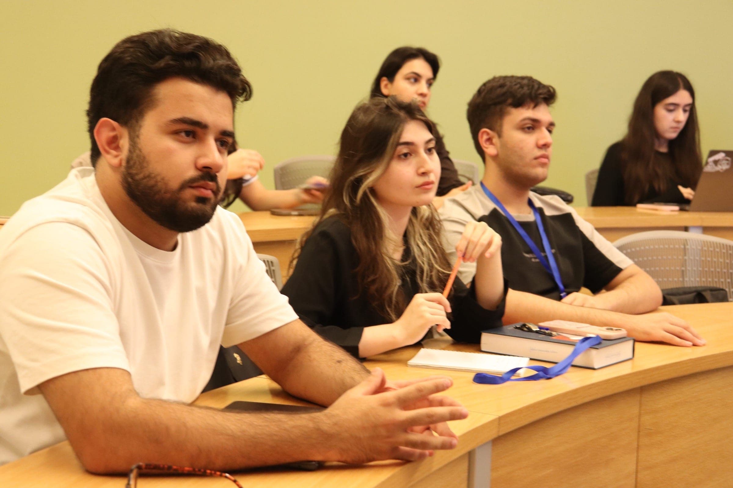 Students at a curved desk following the presentation, notebooks and pens in front of them.