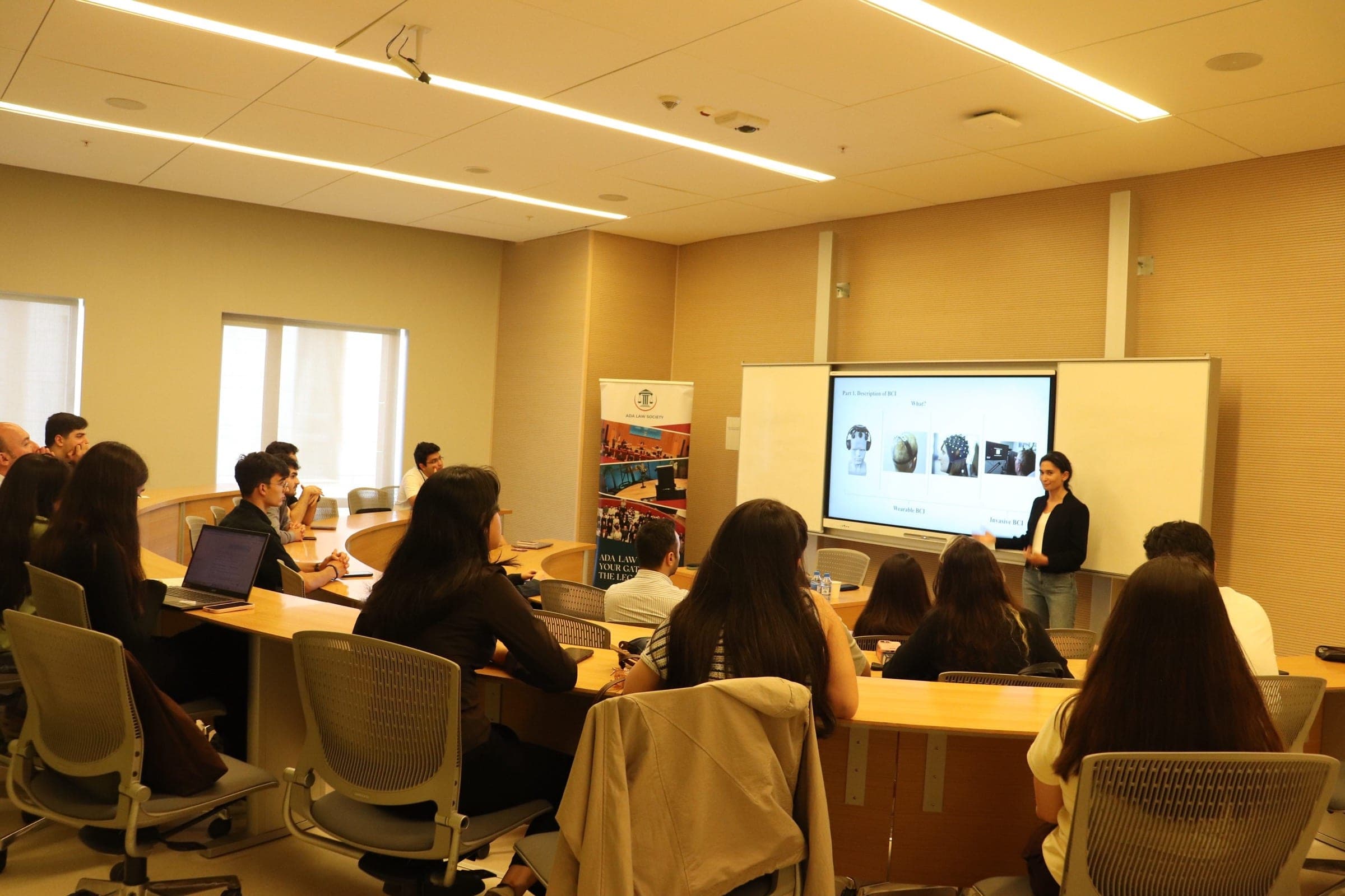 A seminar room seen from the back: students at curved desks facing a screen, with the speaker standing beside it.