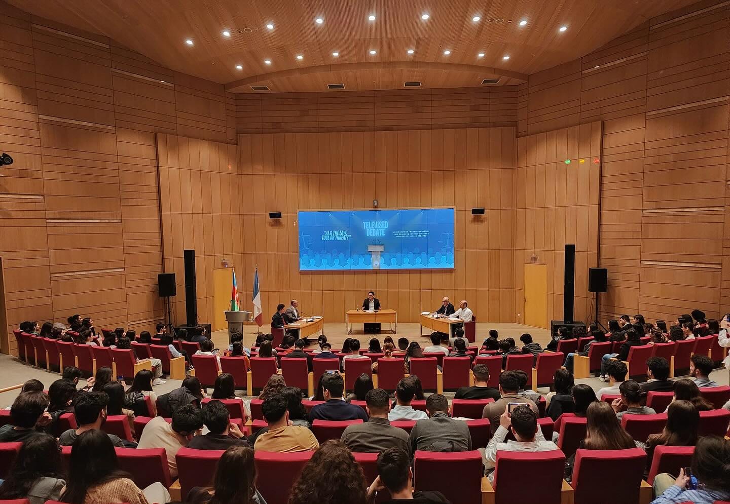 A full wood-panelled auditorium seen from the back rows, a screen lit at the front.