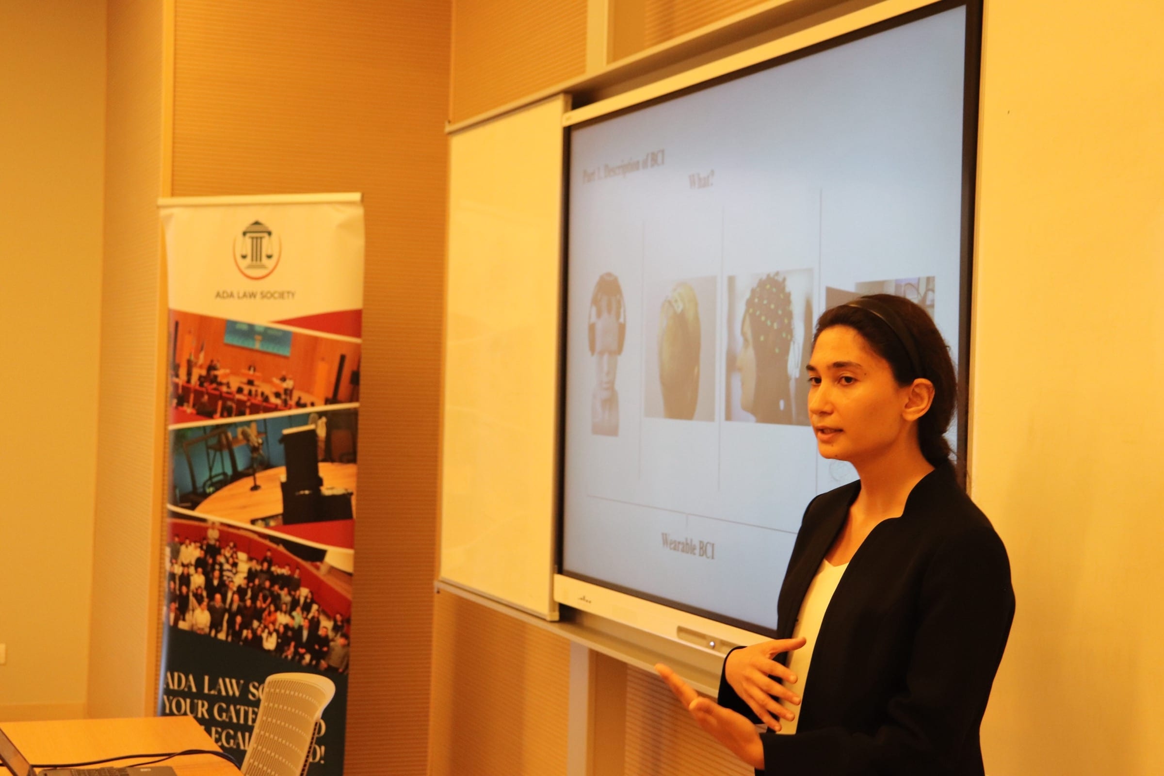 A speaker presenting beside a screen of slides, with an ADA Law Society roll-up banner standing to the left.