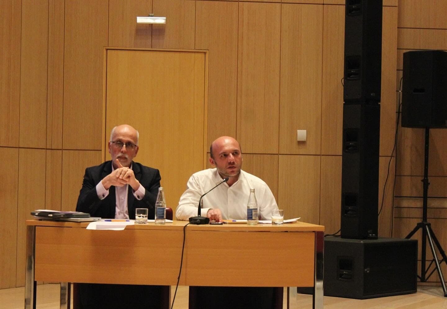 Two participants seated at a table on the stage, speaking into microphones.