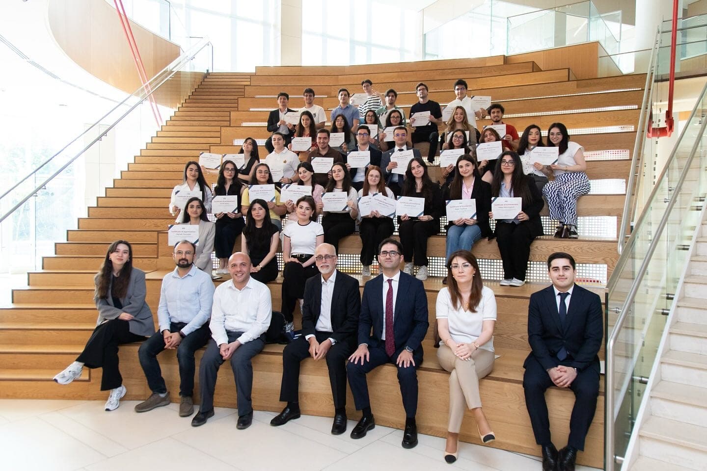 A large group of students seated and standing on a wide staircase, many holding certificates.
