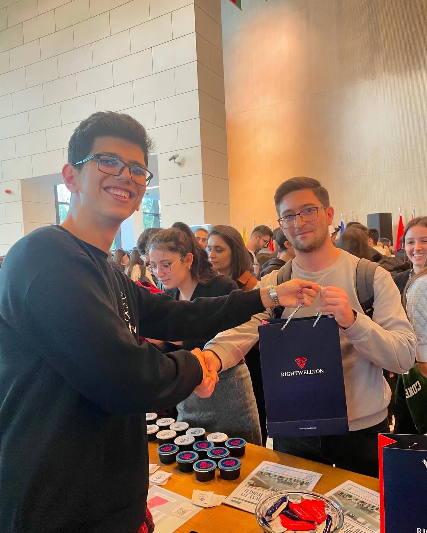 At a busy stand, a student hands a visitor a gift bag printed with the name Rightwellton; other students crowd around behind them.