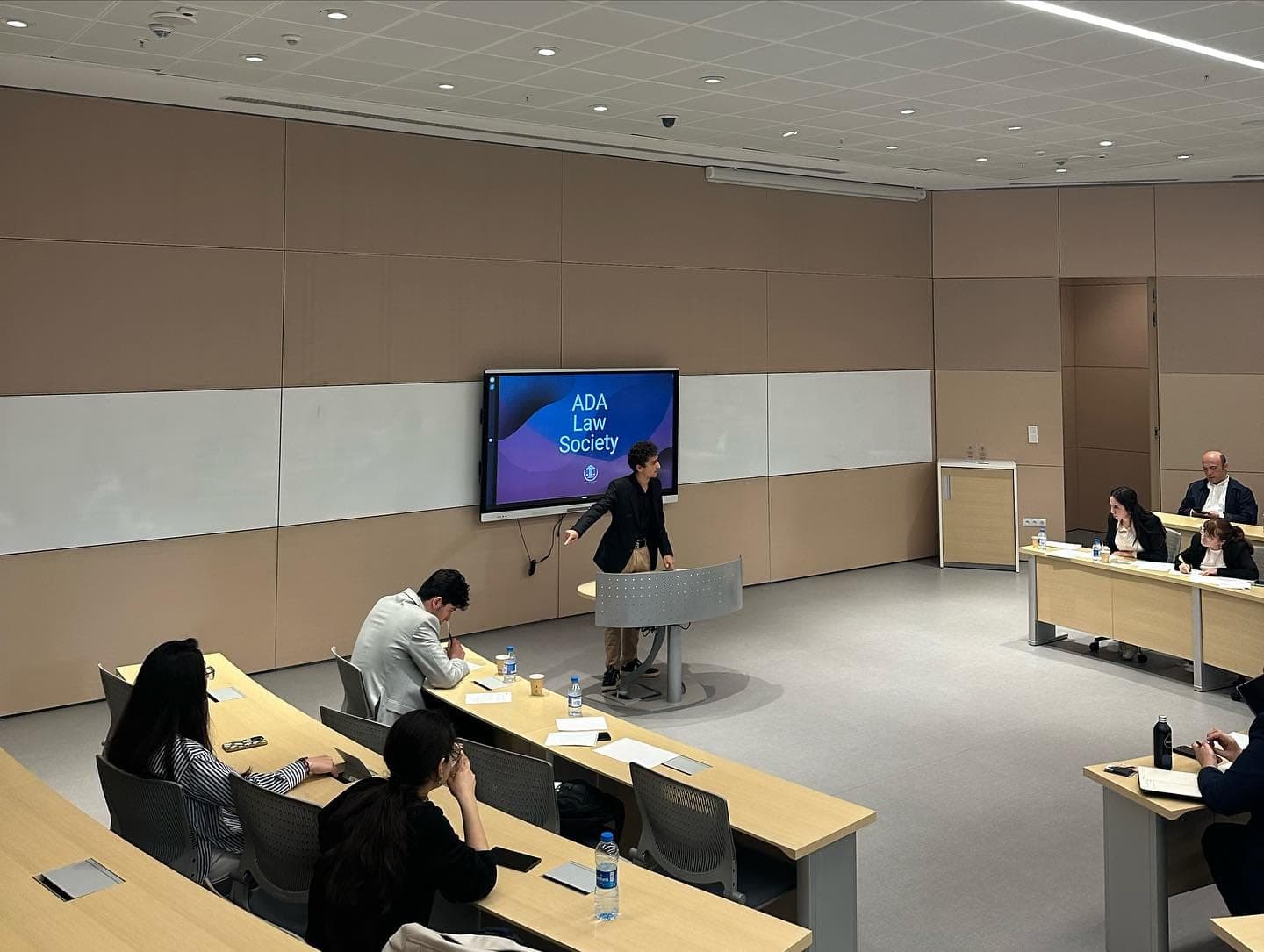 A speaker at a lectern turns towards the screen reading “ADA Law Society”, with participants seated at desks on either side of the floor.