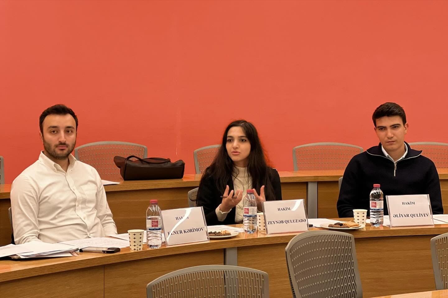 Three participants seated side by side at a table with name placards and notes, in front of a red wall.