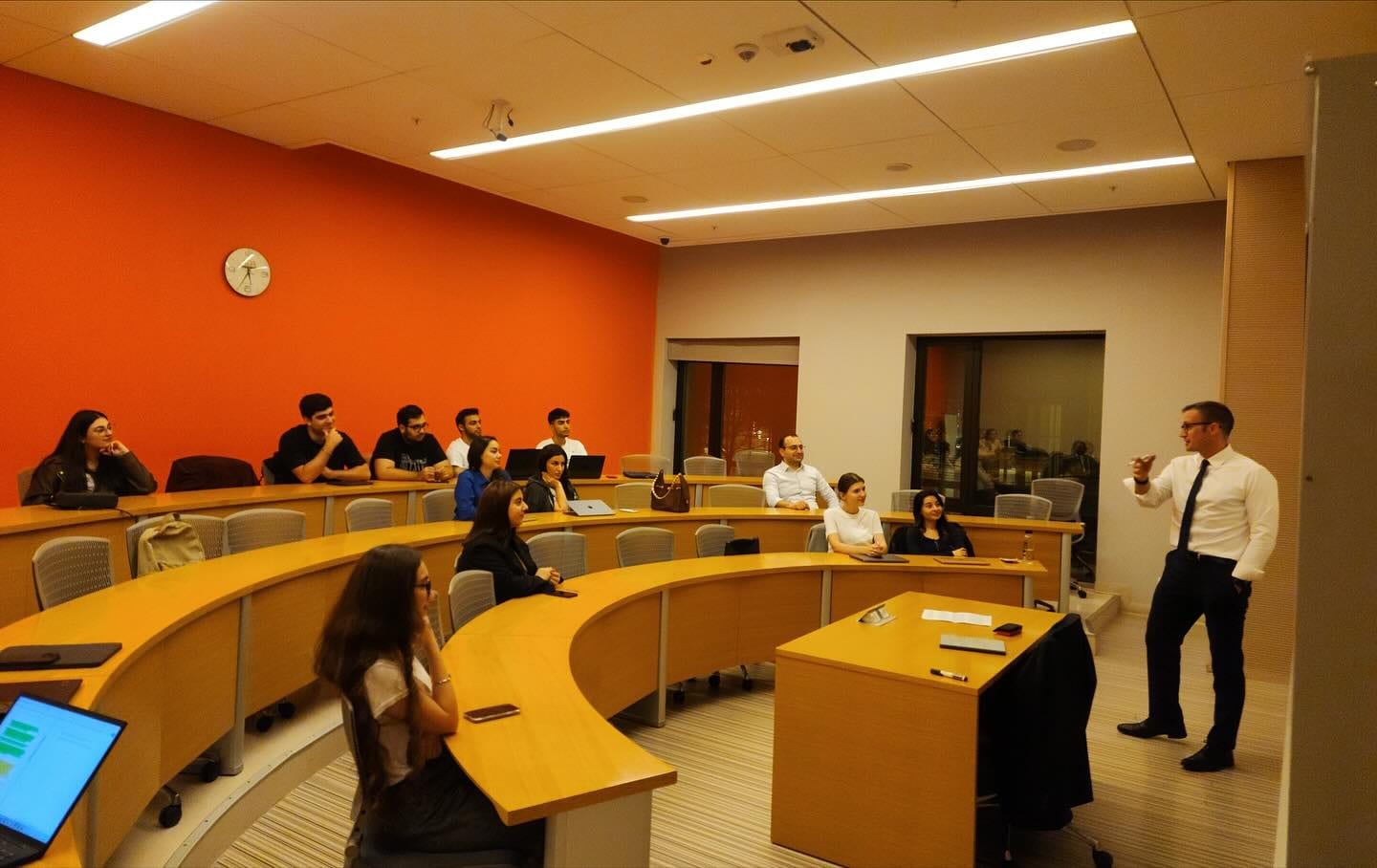 A guest speaker standing at the front of a tiered seminar room with an orange wall, students listening from the curved desks.