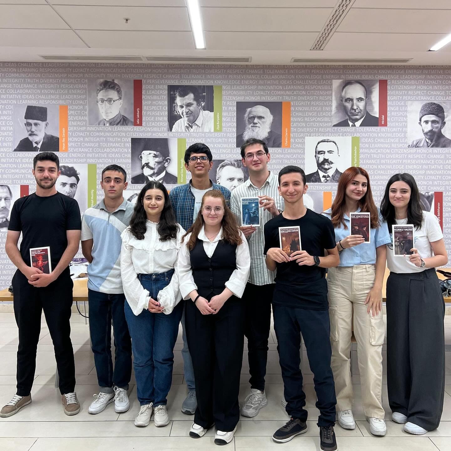 Quiz participants standing in a row, several holding books, in front of a wall of historic portraits.