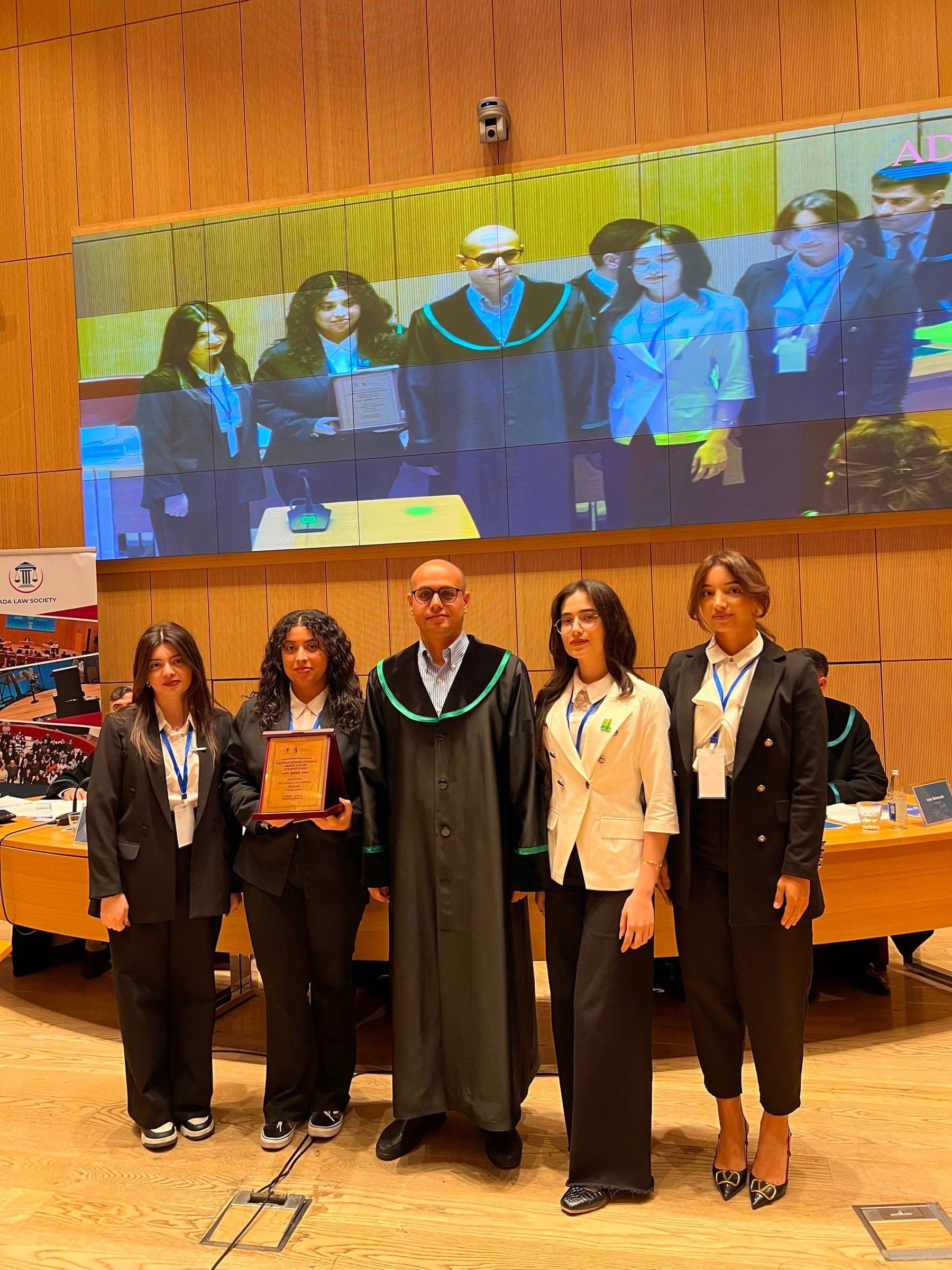 Four participants in formal black standing with a judge in robes who holds a competition plaque, the chamber screen behind them.