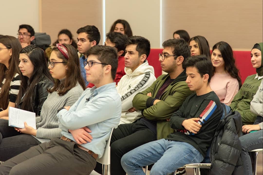 A room full of students seated close together, listening.